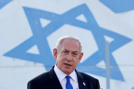 Netanyahu, in giacca e cravatta, parla davanti a una grande bandiera israeliana con il simbolo della Stella di Davide.