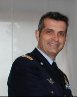 Uomo in uniforme militare IDF scura con insegne, in piedi al chiuso e sorridente alla telecamera.
