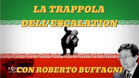 Lo sconcertante discorso alla nazione di Trump Un uomo rannicchiato sotto una grande scritta su una bandiera iraniana, che evoca un discorso alla nazione, con il ritratto di un altro uomo di fronte.
